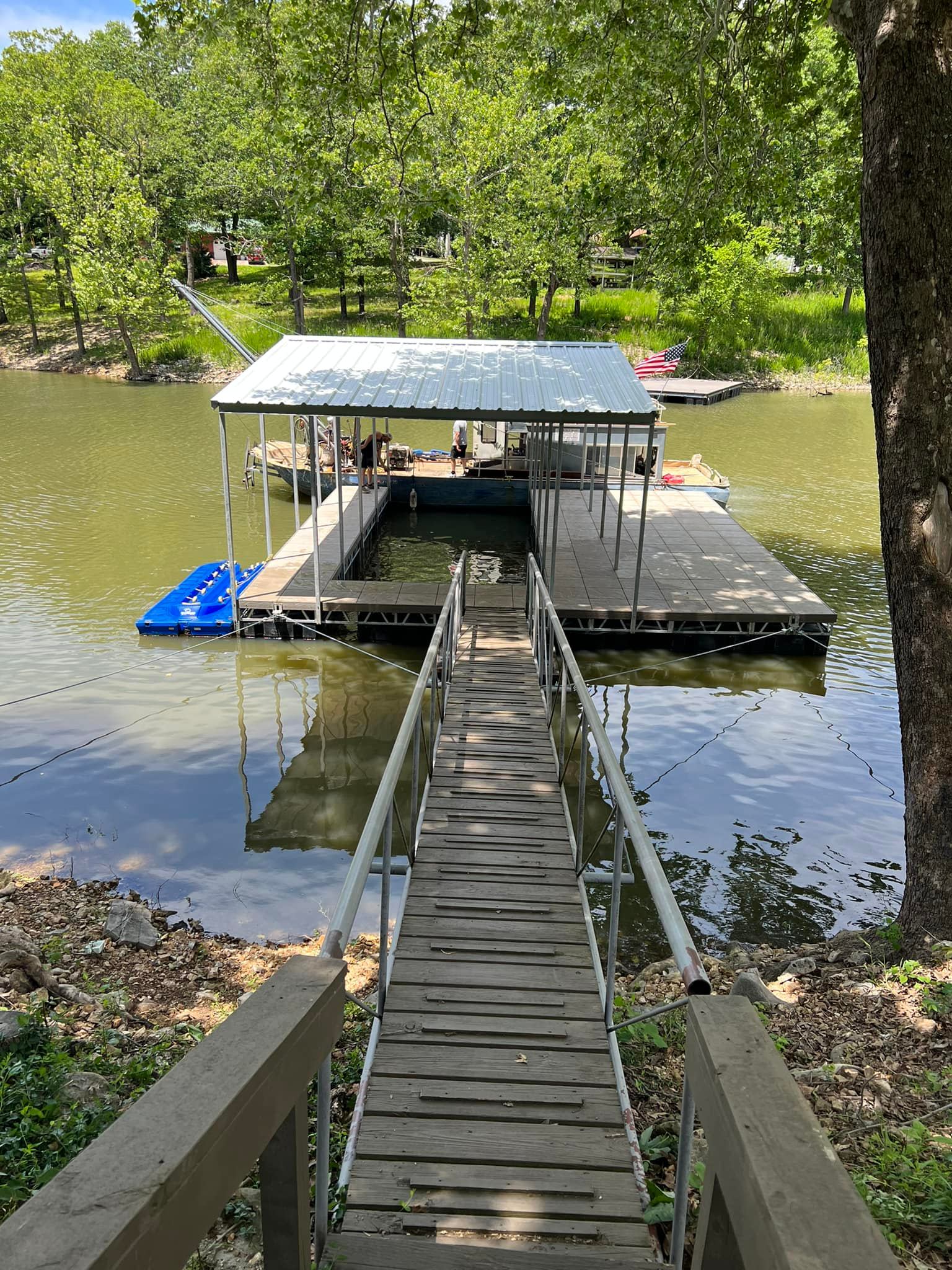 Wooden walkway leading to a covered floating boat dock on Grand Lake in Oklahoma, featuring a metal roof, slip style mooring area, and calm shoreline surroun...