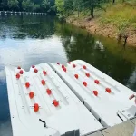 White dual boat lift platforms with red rollers beside a dock on Grand Lake in Oklahoma, shown against a calm wooded shoreline